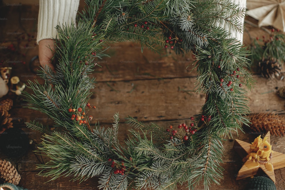 Hands holding stylish rustic wreath on rustic wooden background with pine cones, scissors, candles, ornaments. Top view. Happy holidays. Festive winter workshop. Making xmas wreath