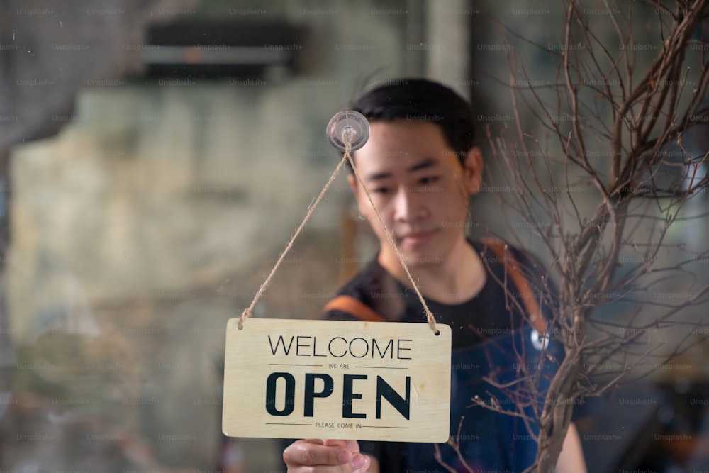 Asian man coffee shop barista walking to cafe door and turning hanging closed sign to open. Male waiter preparing restaurant service to customer. Small business owner and part time job working concept