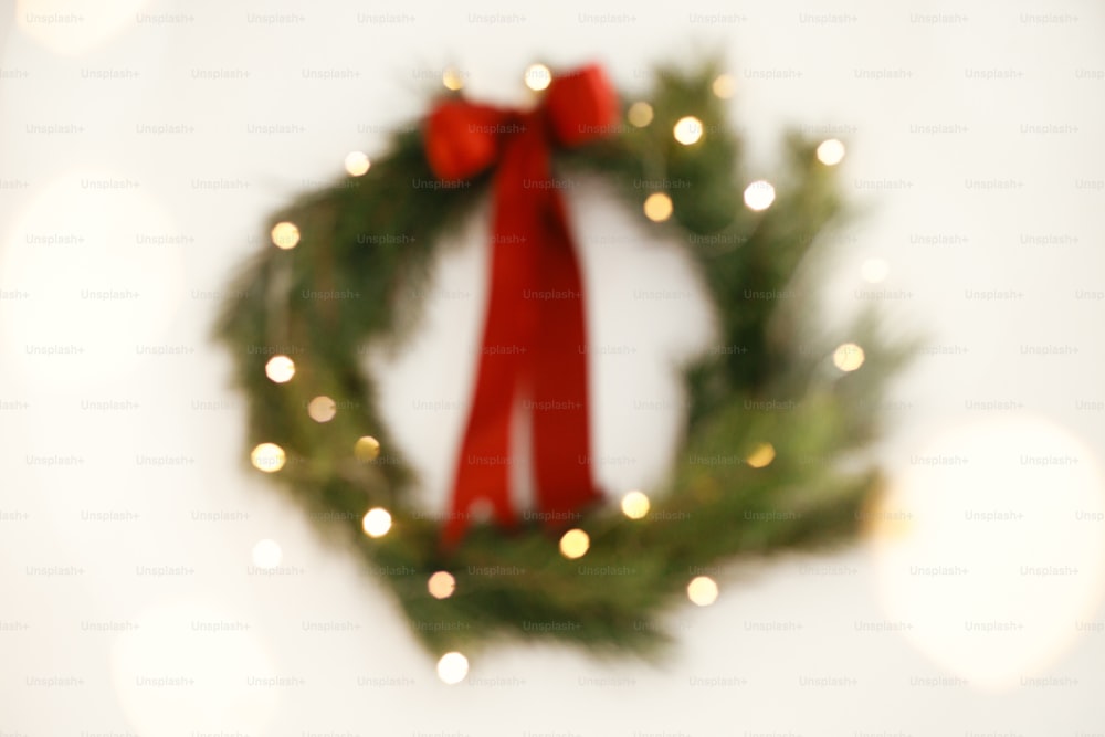 Weihnachten abstrakter Hintergrund. Unscharfer stilvoller Weihnachtskranz mit roter Schleife und goldenen Lichtern an weißer Wand. Frohe Weihnachten! Traditioneller Weihnachtskranz mit Tannenzweigen und rotem Band