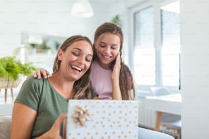 Young woman and girl at home celebrating mother's day sitting on sofa daughter hugging mother kissing cheek mom laughing joyful holding gift box