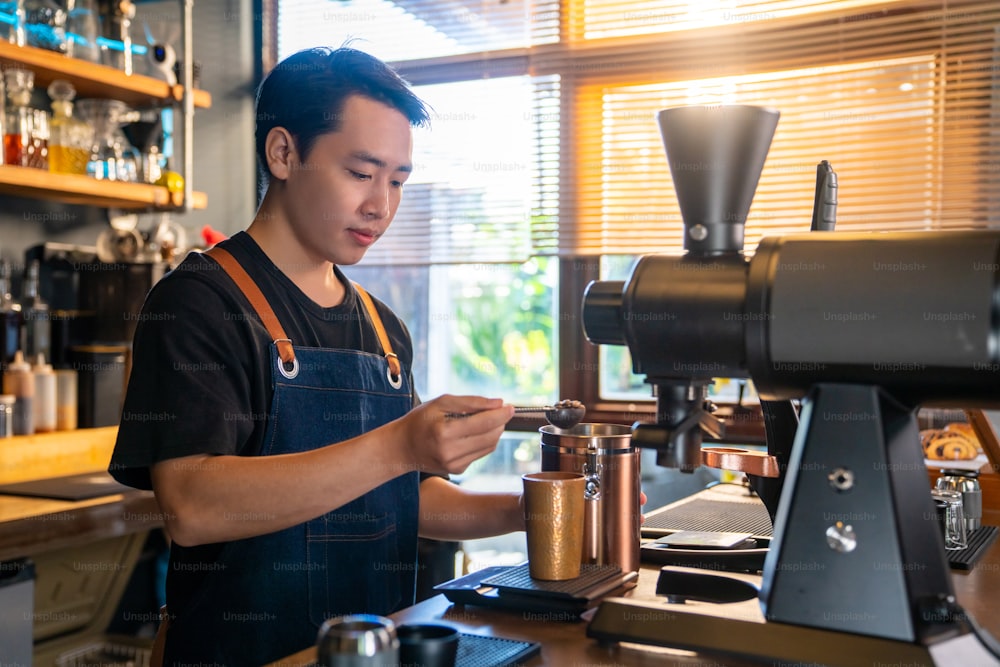 カフェのカウンターバーで焙煎したコーヒー豆を挽くコーヒーグラインダーマシンを使用するアジア人男性バリスタ。客にブラックコーヒーを淹れるコーヒーショップの男性店主。スモールビジネスレストランの食べ物と飲み物のコンセプト