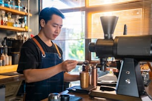 Asian man barista using coffee grinder machine grinding roasted coffee beans on counter bar at cafe. Male coffee shop owner brewing black coffee serving to customer. Small business restaurant food and drink concept