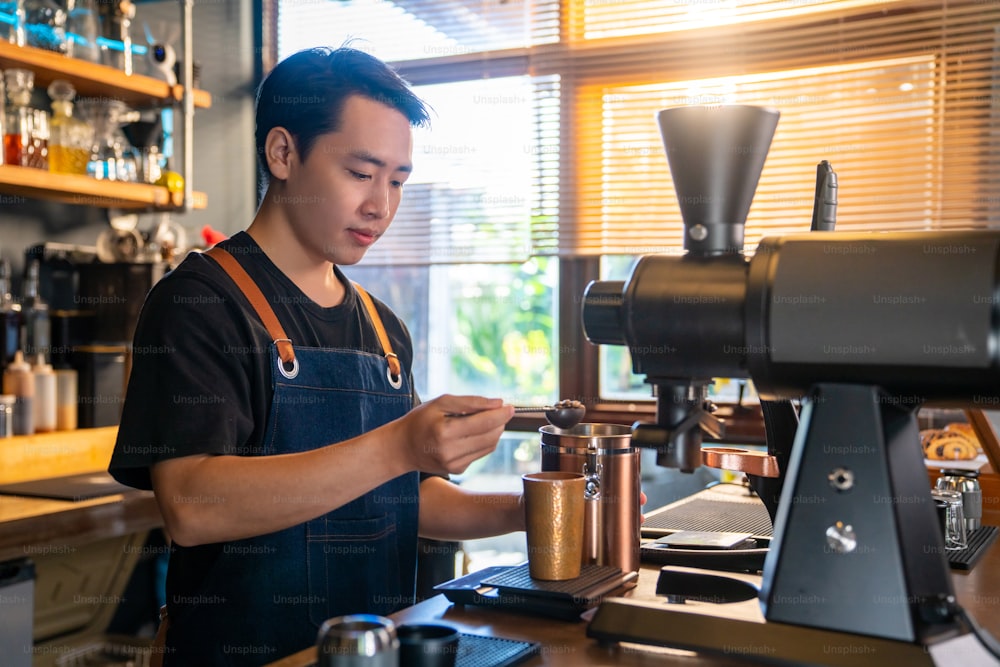 Asian man barista using coffee grinder machine grinding roasted coffee beans on counter bar at cafe. Male coffee shop owner brewing black coffee serving to customer. Small business restaurant food and drink concept