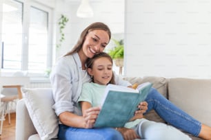 Loving young mother reading book to adorable little daughter, sitting on cozy sofa in living room, mum teaching preschool girl child, family spending weekend at home together, children education