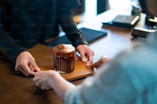 Asian woman barista serving iced cocoa with froth milk in the glass to customer on bar counter at cafe. Female coffee shop waitress employee taking order from client. Small business coffee shop owner concept