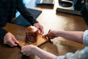 Asian woman barista serving iced cocoa with froth milk in the glass to customer on bar counter at cafe. Female coffee shop waitress employee taking order from client. Small business coffee shop owner concept
