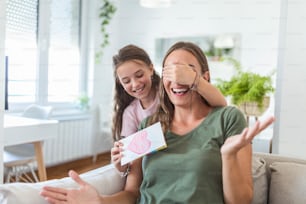 Happy little girl congratulating smiling mother and giving card with red heart during holiday celebration at home, covering her eyes and surprising her