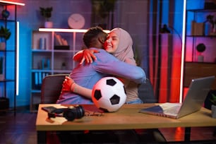 Mujer árabe feliz en hiyab abrazando a un hombre afroamericano mientras mira un partido de fútbol en una computadora portátil. Pareja joven celebrando la victoria del equipo favorito. Concepto de apoyo y emociones.