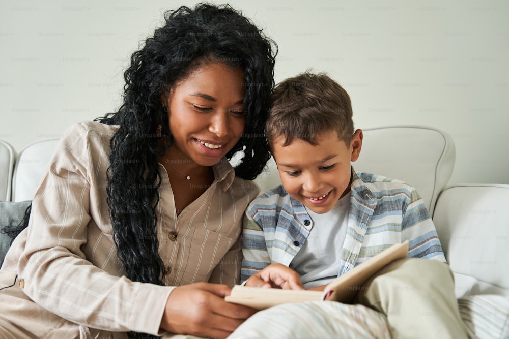 Madre negra leyendo un libro con su hijo en el sofá. Concepto de relaciones familiares y crianza de los hijos. Idea de maternidad. Gente sonriente en el interior de la sala de estar