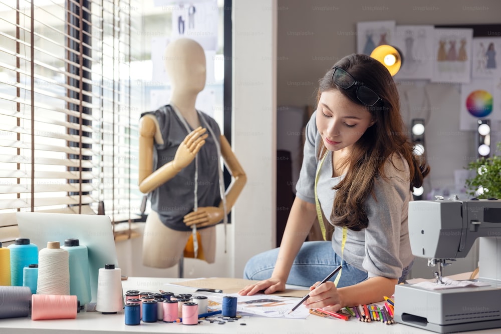While an assessment of performance at clothes in the background, a professional tailor designer makes notes and drawings in a notebook, Business Owner With Tape Measure Working In Fashion Sitting On Desk