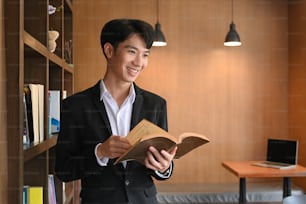 Lächelnder Geschäftsmann, der ein Buch hält und an einem modernen Arbeitsplatz steht.