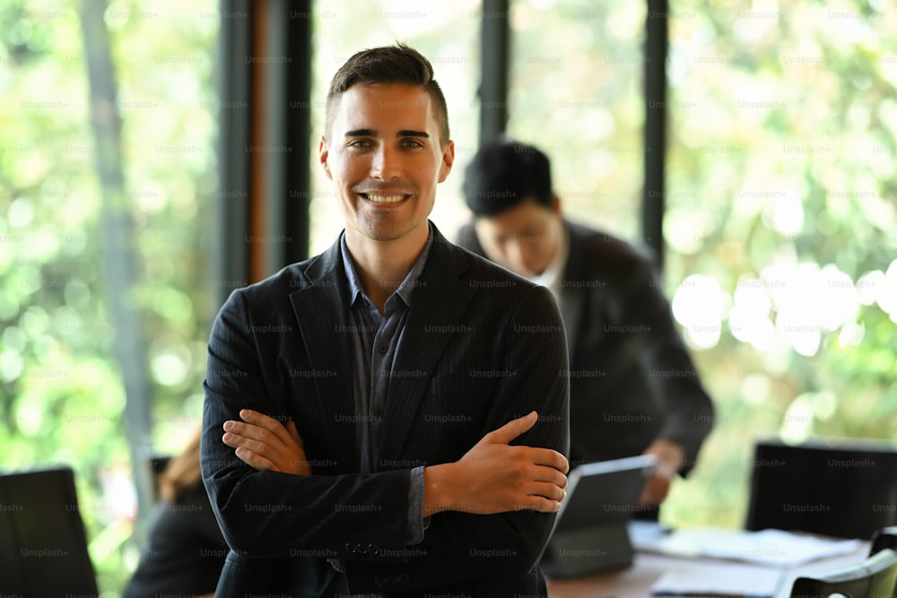Handsome businessman standing with crossed arms and group of colleagues in the background.