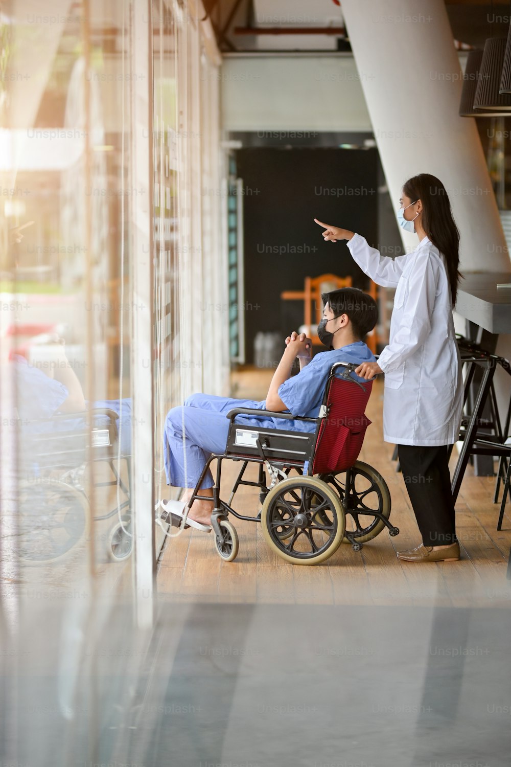 A young male patient in a wheelchair sits in a hospital corridor against a large window, accompanied by a female doctor.