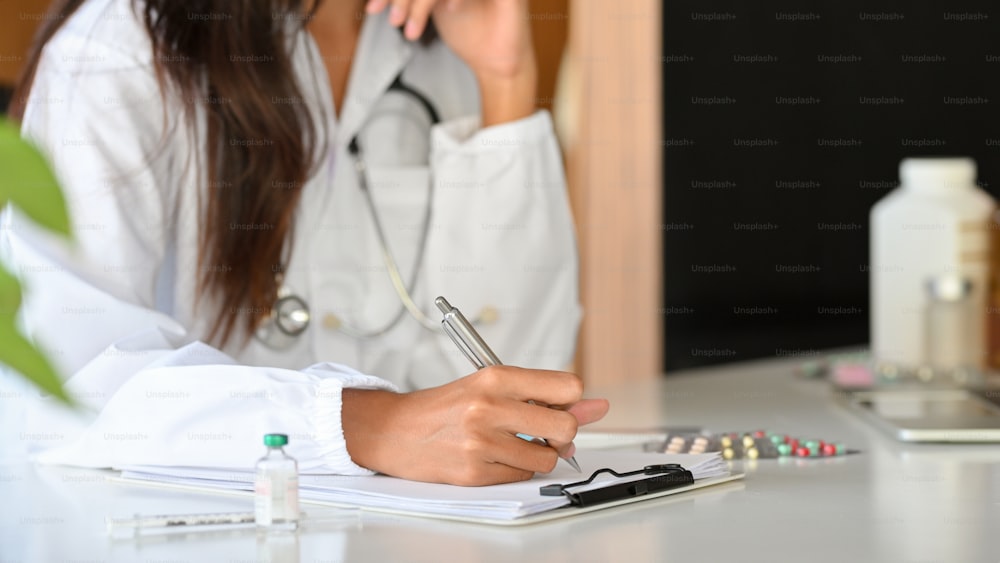 女性の開業医や薬剤師が薬の記録クリップボードに処方箋を書いている様子を撮影した写真。机の上のワクチンバイアル、錠剤、ビタミンボトル