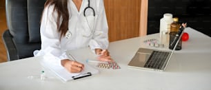 Abgeschnittenes Bild einer Ärztin oder Apothekerin, die ein medizinisches Formular ausfüllt, während sie sich ein medizinisches Online-Webinar auf ihrem Laptop im Krankenhausbüro ansieht.