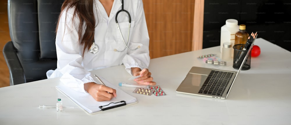 Imagem recortada de uma médica ou farmacêutica preenchendo um formulário médico enquanto assistia a um webinar médico on-line em seu laptop no consultório do hospital.