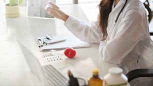 Side view of Asian female doctor working on her desk. laptop, pills, medical clipboard, red heart, glasses on desk