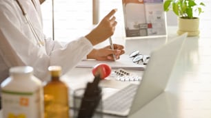 Pharmacist is researching medical drugs and writing medical prescriptions. On the work table, there are pills, vitamin bottles, a laptop, and glasses. Cropped image