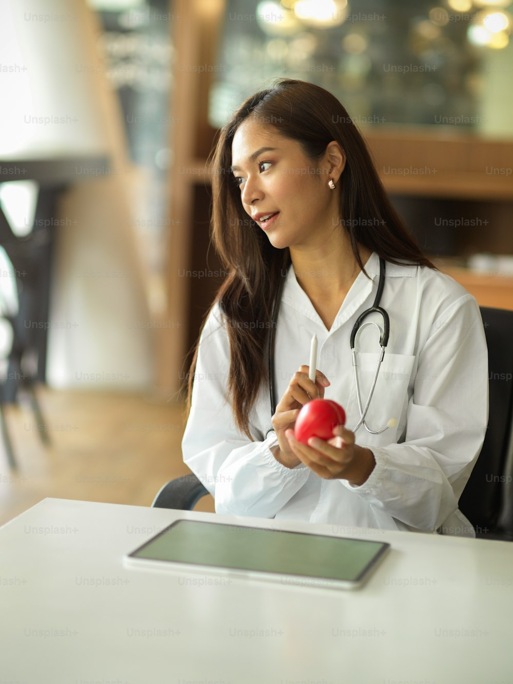 Portrait of a beautiful female doctor or physician wearing a white gown and stethoscope, sits at her desk while holding a red heart.