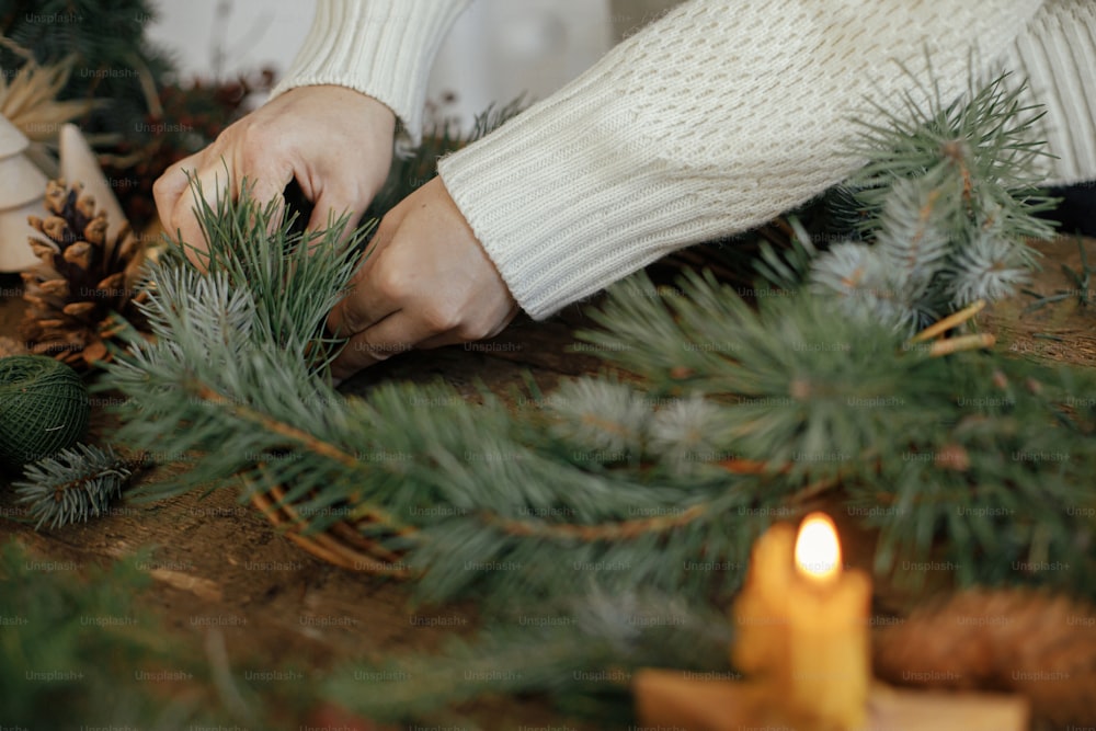 Making rustic christmas wreath close up. Woman hands holding fir branch and arranging christmas wreath on rustic wooden background with candle, pine cones, thread. Winter holiday preparations