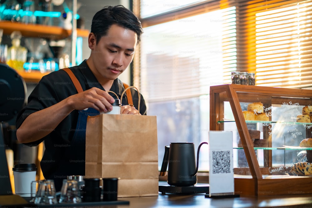 Confidence Asian man waiter or barista working at cafe. Male coffee shop owner preparing takeaway order hot coffee to customer on bar counter. Small business entrepreneur food and drink concept.