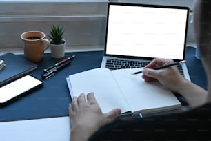 Young man working online with laptop computer and writing information on notebook.