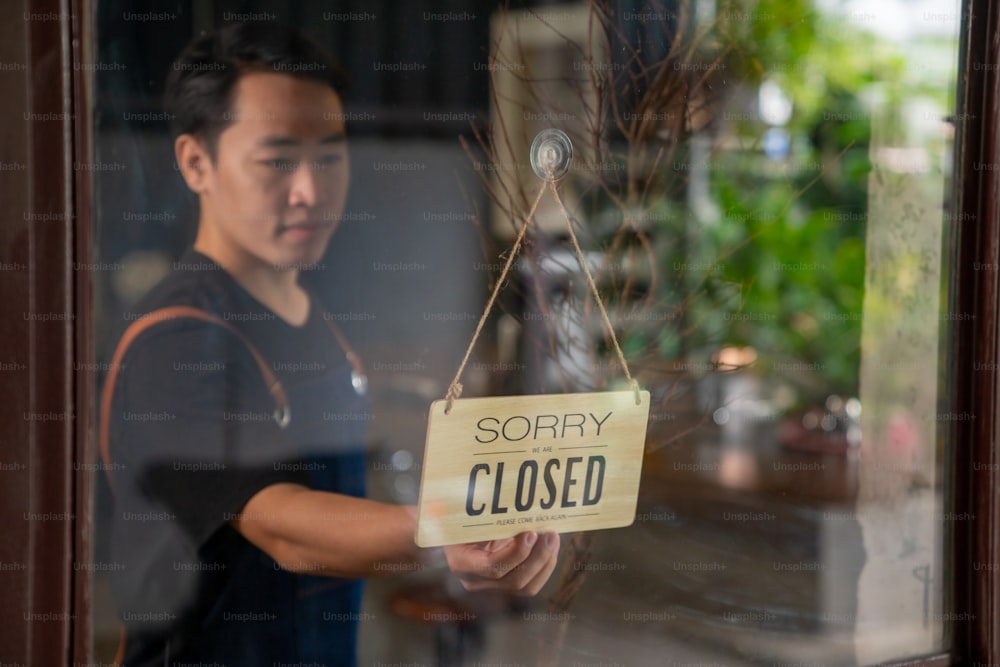 Asian man coffee shop barista walking to cafe door and turning hanging closed sign to open. Male waiter preparing restaurant service to customer. Small business owner and part time job working concept
