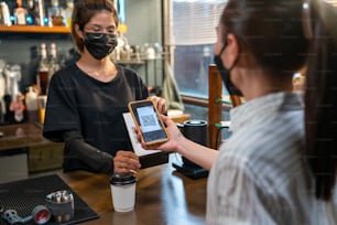Una mujer asiática usa una máscara facial protectora durante la pandemia de COVID-19 usando un código de barras de escaneo de teléfonos inteligentes para realizar pagos sin contacto en una cafetería. Pequeña empresa con concepto de pago electrónico de banca móvil