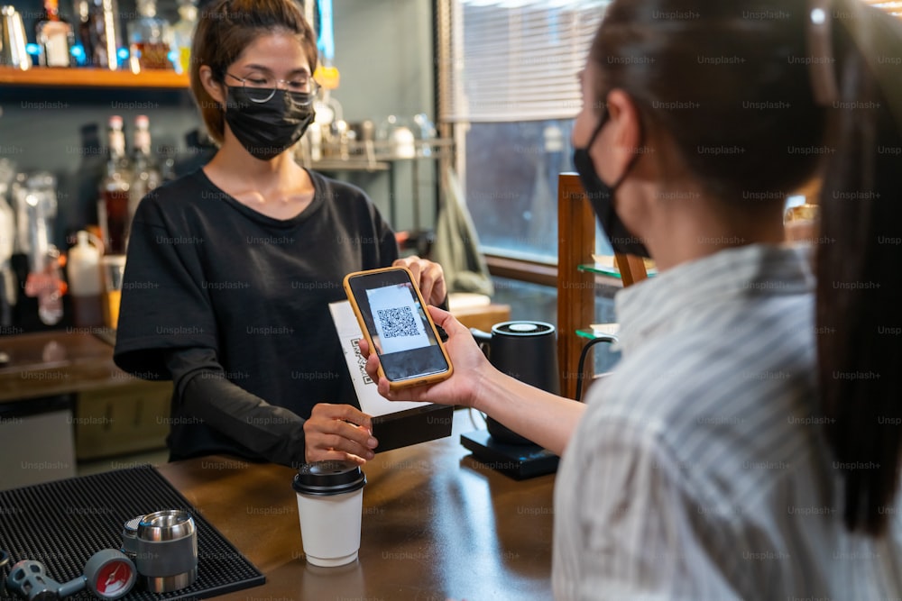Asian woman wear protective face mask during COVID-19 pandemic using smartphone scanning bar code make contactless payment at coffee shop. Small business with mobile banking electronic payment concept
