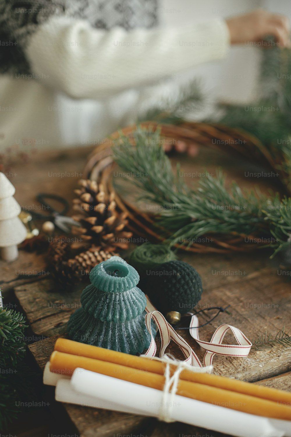 Elegante vela festiva de Navidad sobre fondo de mujer en suéter acogedor que hace corona navideña sobre fondo de madera rústica. Espacio para el texto. Felices Fiestas. Taller festivo de invierno