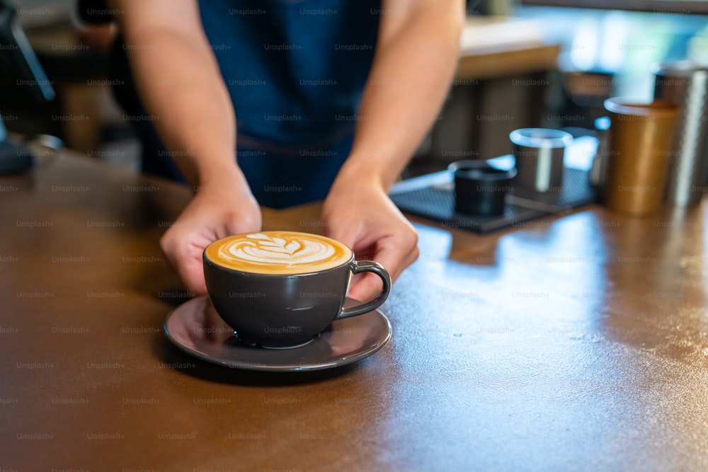 Asian man barista making hot coffee latte in coffee cup to customer on bar counter at cafe. Male coffee shop waiter serving hot coffee with milk to client. Small business restaurant owner concept.