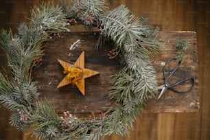 Feliz Navidad y Felices Fiestas. Elegante corona navideña con ramas de abeto y bayas, vela estrella y tijeras sobre fondo rústico de madera.  Taller festivo de invierno. Tendido plano