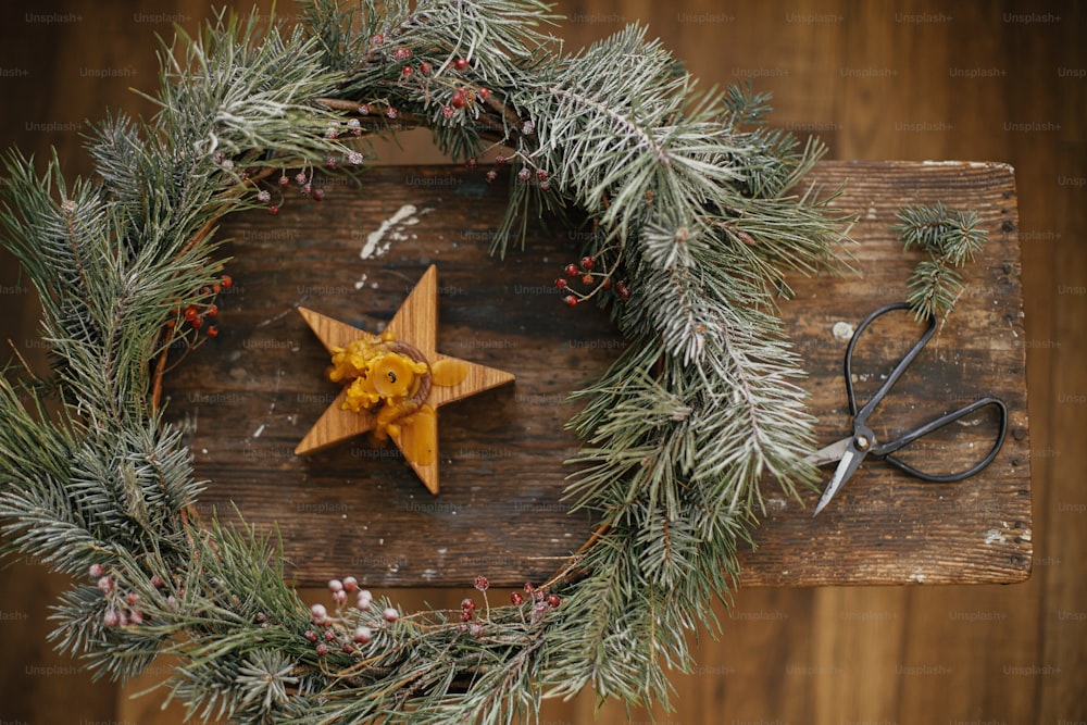 Joyeux Noël et Joyeuses Fêtes. Couronne de Noël élégante avec des branches d’épinette et des baies, une bougie étoile et des ciseaux sur fond en bois rustique.  Atelier d’hiver festif. Pose à plat