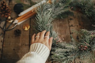 Hand with stylish rings on rustic christmas wreath with pine branches and red berries in snow on rustic old wooden background close up. Happy holidays. Making rustic xmas wreath, holiday workshop