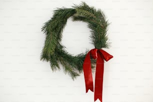 Couronne de Noël élégante avec noeud rouge accroché sur le mur blanc. Joyeux Noël et Joyeuses Fêtes ! Couronne de Noël traditionnelle simple avec des branches de pin et un ruban rouge isolé sur fond blanc
