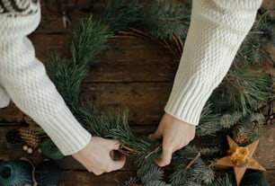 Woman hands holding pine branches and arranging christmas wreath on rustic wooden background with pine cones, scissors, candle. Top view. Winter holiday preparations. Festive workshop