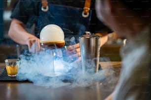 Asian man barista shaking iced black coffee in shaker and pouring in cocktail glass on bar counter at coffee shop. Male coffee shop owner serving cold drink coffee to customer. Small business restaurant concept.