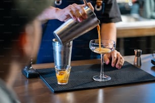 Asian man barista shaking iced black coffee in shaker and pouring in cocktail glass on bar counter at coffee shop. Male coffee shop owner serving cold drink coffee to customer. Small business restaurant concept.