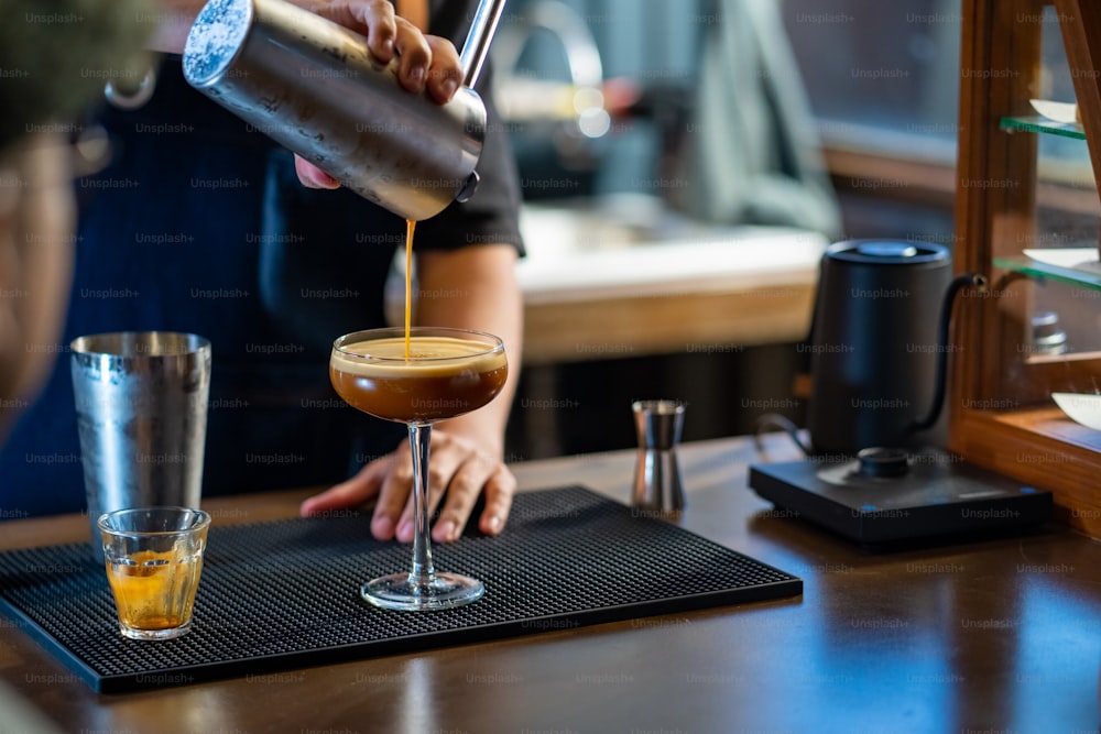 Asian man barista shaking iced black coffee in shaker and pouring in cocktail glass on bar counter at coffee shop. Male coffee shop owner serving cold drink coffee to customer. Small business restaurant concept.