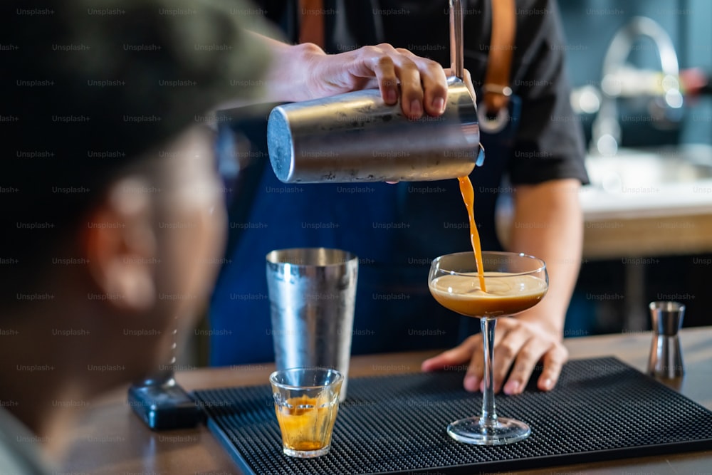 Asian man barista shaking iced black coffee in shaker and pouring in cocktail glass on bar counter at coffee shop. Male coffee shop owner serving cold drink coffee to customer. Small business restaurant concept.