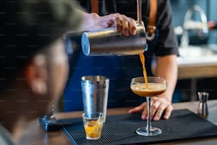 Asian man barista shaking iced black coffee in shaker and pouring in cocktail glass on bar counter at coffee shop. Male coffee shop owner serving cold drink coffee to customer. Small business restaurant concept.