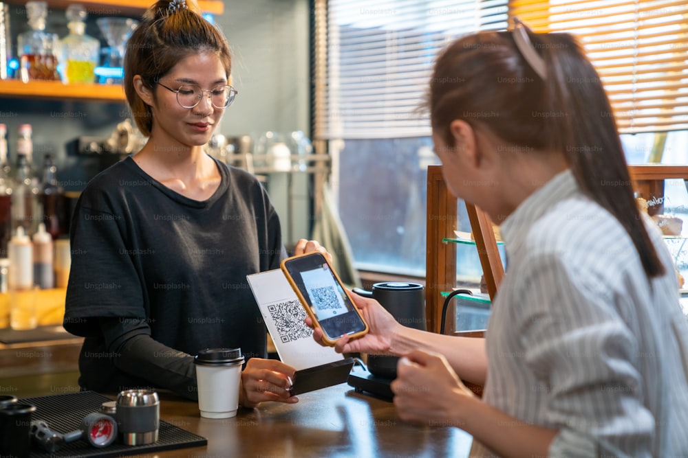 Modern Asian woman using smartphone scanning bar code making contactless payment checkout her coffee at coffee shop. Small business owner with electronic mobile banking contactless payment concept