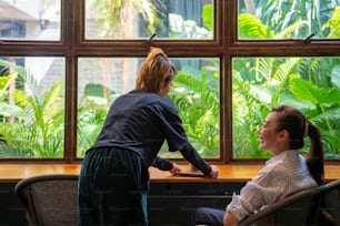 カフェの窓際に座っている客にアイスチョコレートとミルクを振る舞うアジア人女性バリスタ。喫茶店のウェイトレスの女性従業員が客から注文を受ける。スモールビジネスコーヒーショップのオーナーコンセプト