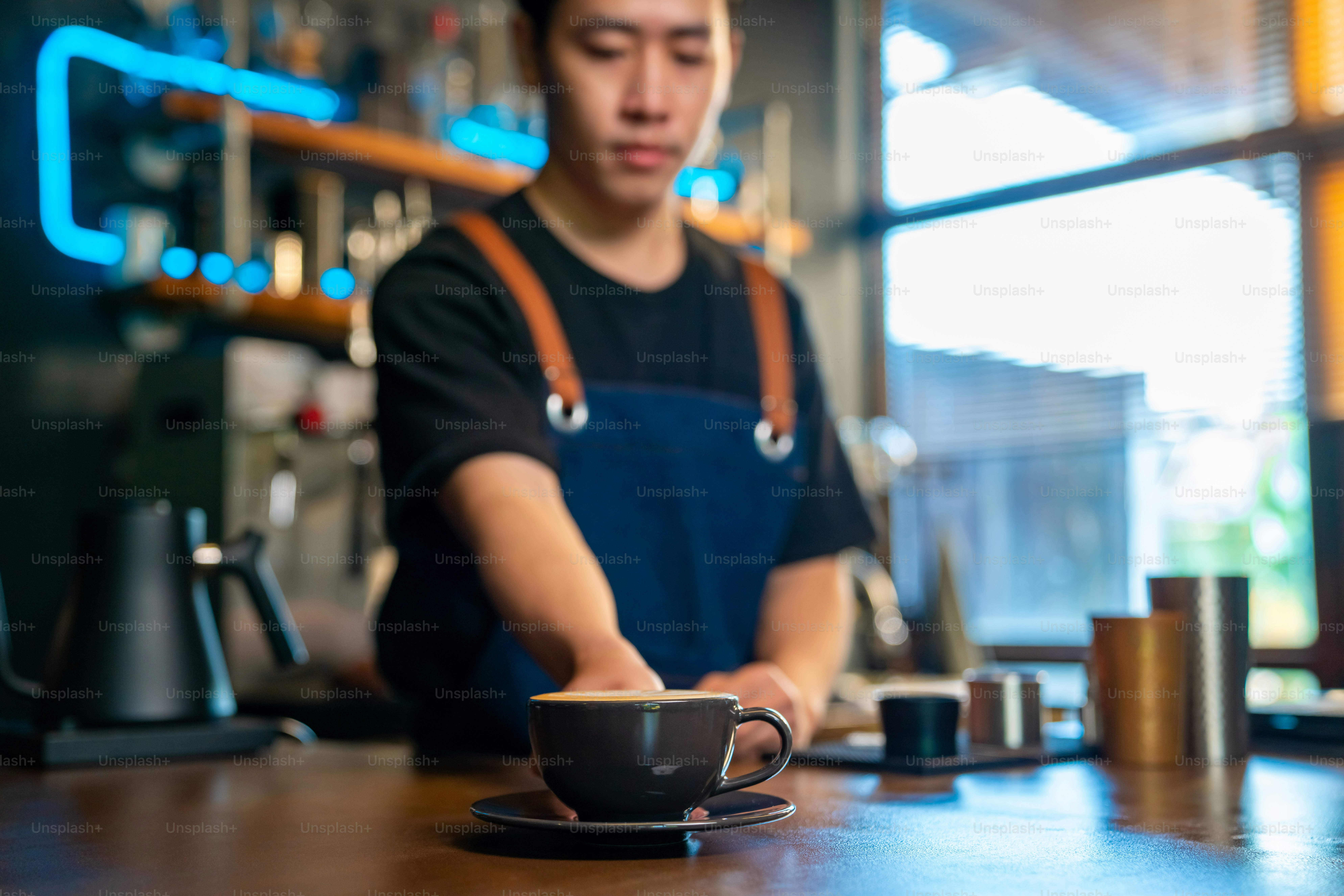 Asian man barista making hot coffee latte in coffee cup to customer on bar counter at cafe. Male coffee shop waiter serving hot coffee with milk to client. Small business restaurant owner concept.