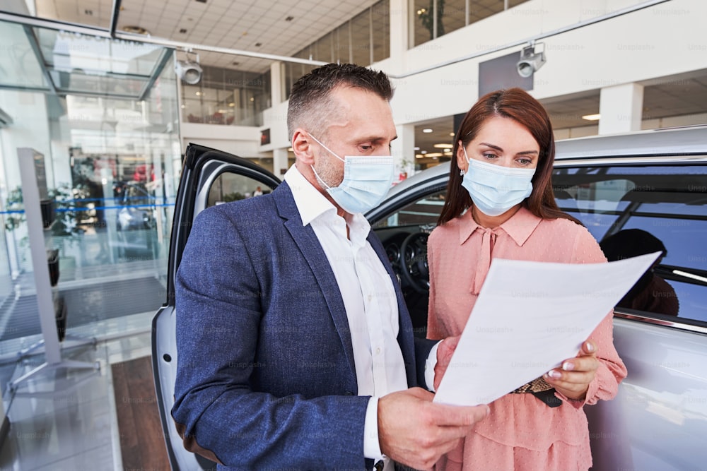 Man and woman having medical masks on their faces looking at the document while standing near the car