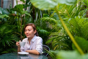 Asiatische Geschäftsfrau, die draußen im Coffeeshop-Garten sitzt, trinkt Kaffee Latte mit Milch in der Kaffeetasse. Selbstbewusste Freiberuflerin, die im Freien im Café arbeitet. Konzept eines kleinen Business-Restaurants.