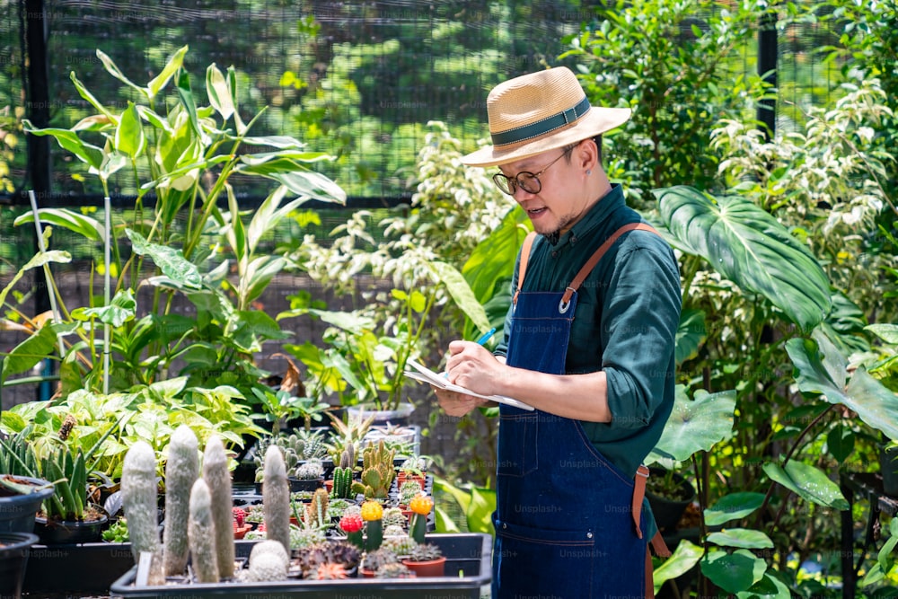 온실 정원에서 관엽 식물과 꽃을 돌보는 아시아 남자 정원사. 남성 식물 상점 주인은 매장에서 식물 주문을 계산하고 확인합니다. 중소 기업 기업가 및 환경 공장 돌보는 개념.