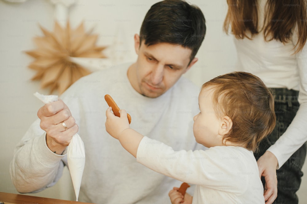 Stylish mother, father and cute little daughter decorating christmas gingerbread cookies with icing in modern room, funny authentic moments. Happy family time together. Xmas holiday preparations