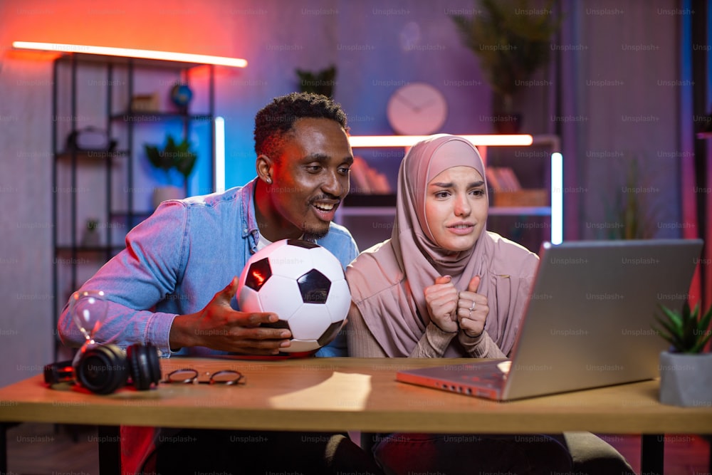 Hombre africano preocupado y mujer musulmana viendo un partido de fútbol en una computadora portátil inalámbrica en casa. Joven pareja multirracial disfrutando del tiempo que pasan juntos. Concepto de emociones y emoción.