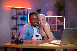 Positive multicultural couple sitting in hugs at table while watching football game on wireless laptop. Entertainment during evening time at home. Soccer fan concept.